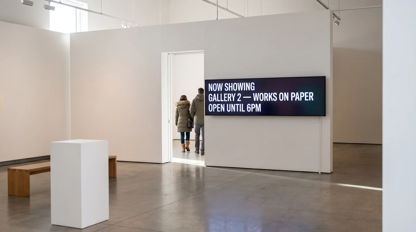 Wide wall-mounted screen in a white-walled art gallery room reading Now showing, Gallery 2 works on paper, Open until 6pm, beside an open doorway with two visitors walking through