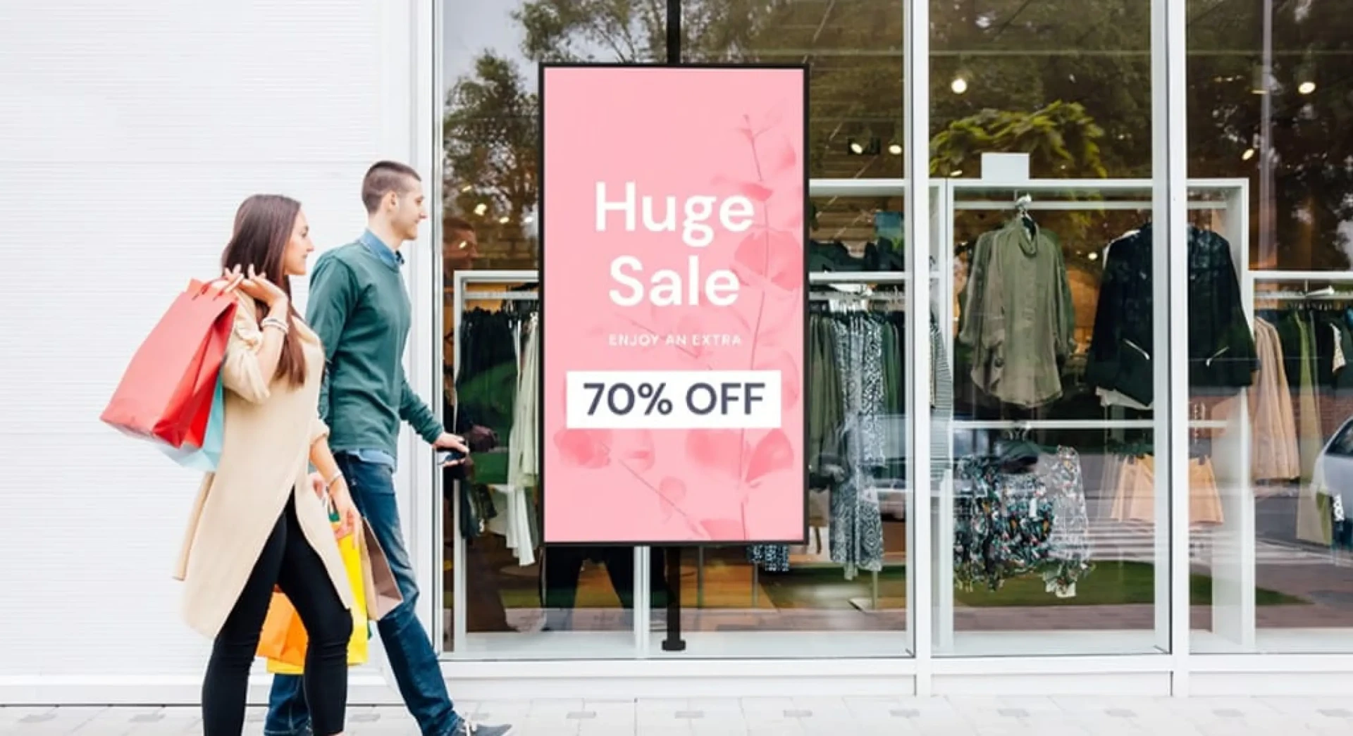Shoppers walking past a clothing store storefront window with a portrait digital poster advertising a Huge Sale at 70% off