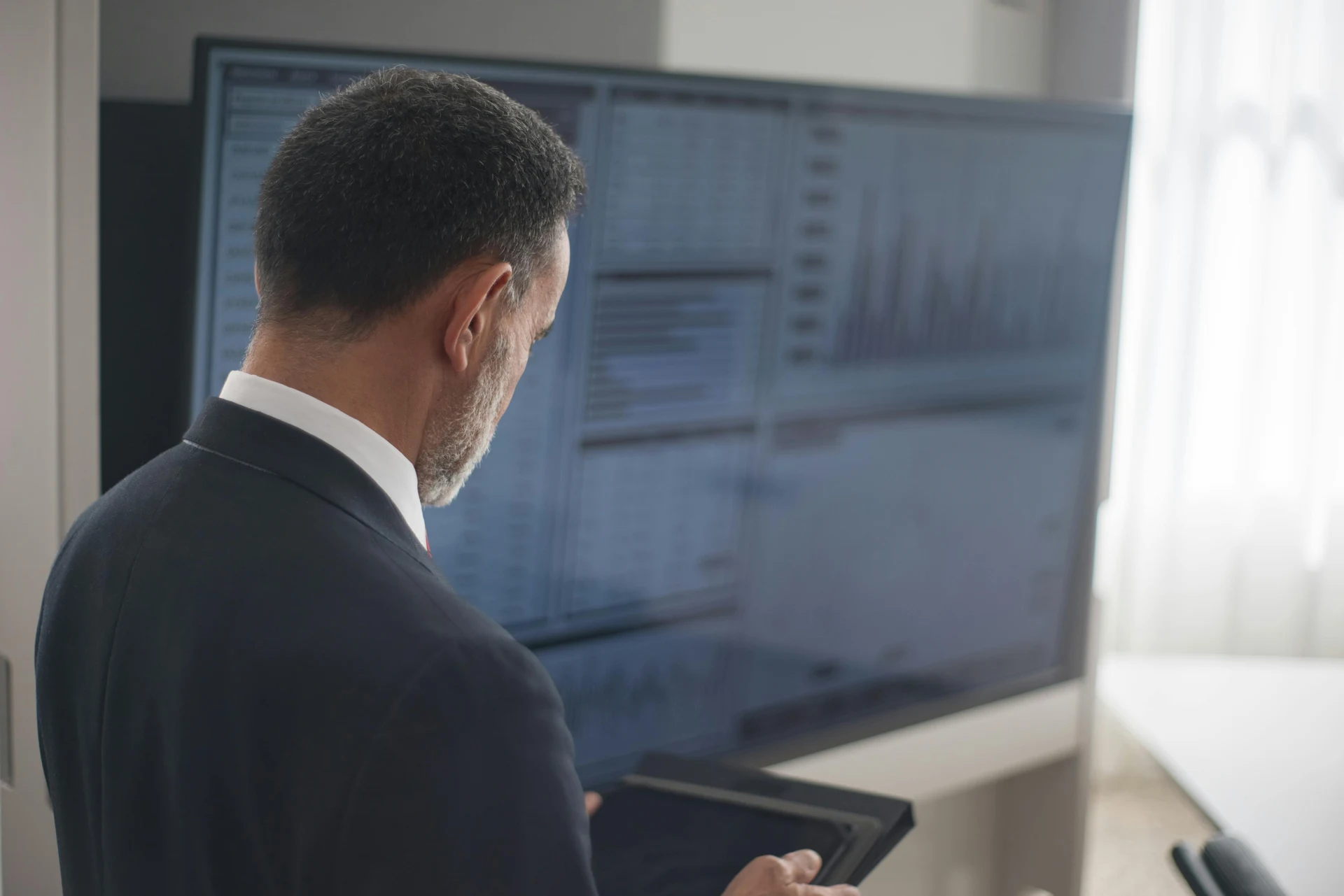 Executive in a suit reviewing a large office display showing revenue, margin, and forecast dashboards while holding a tablet