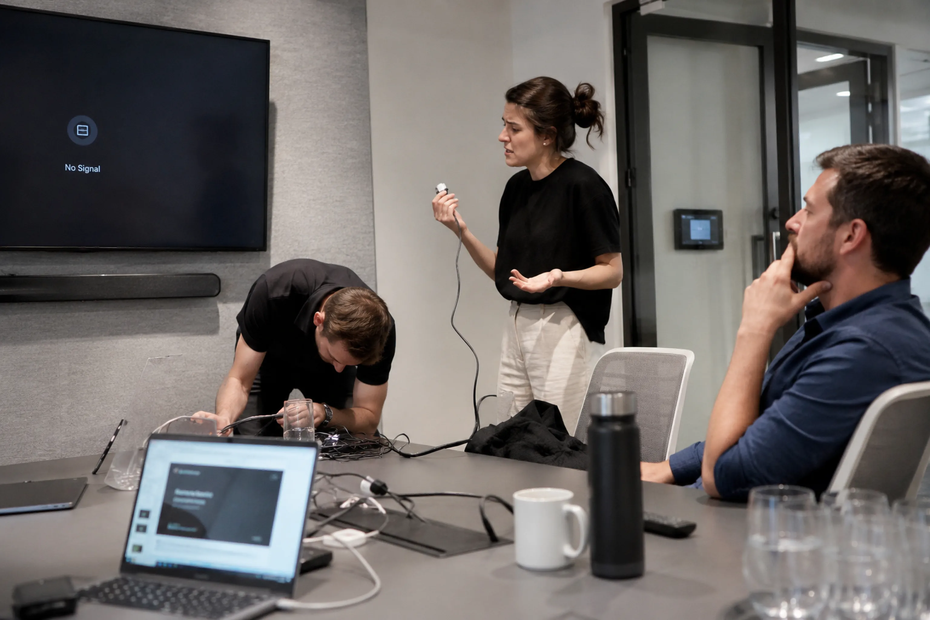 Frustrated meeting attendees fiddling with cables under a "No Signal" meeting room TV