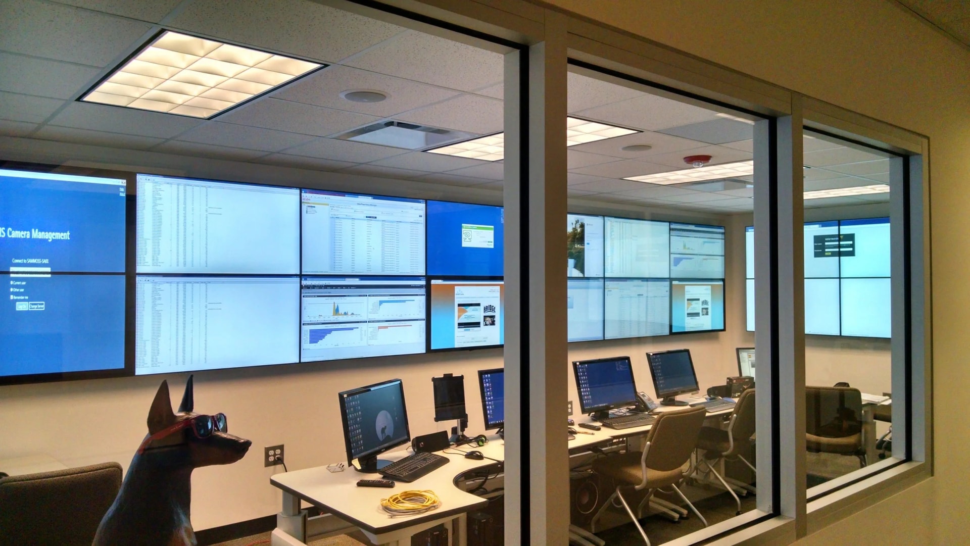 Glass-walled operations room seen from a corridor, with a wall of monitoring screens running unattended
