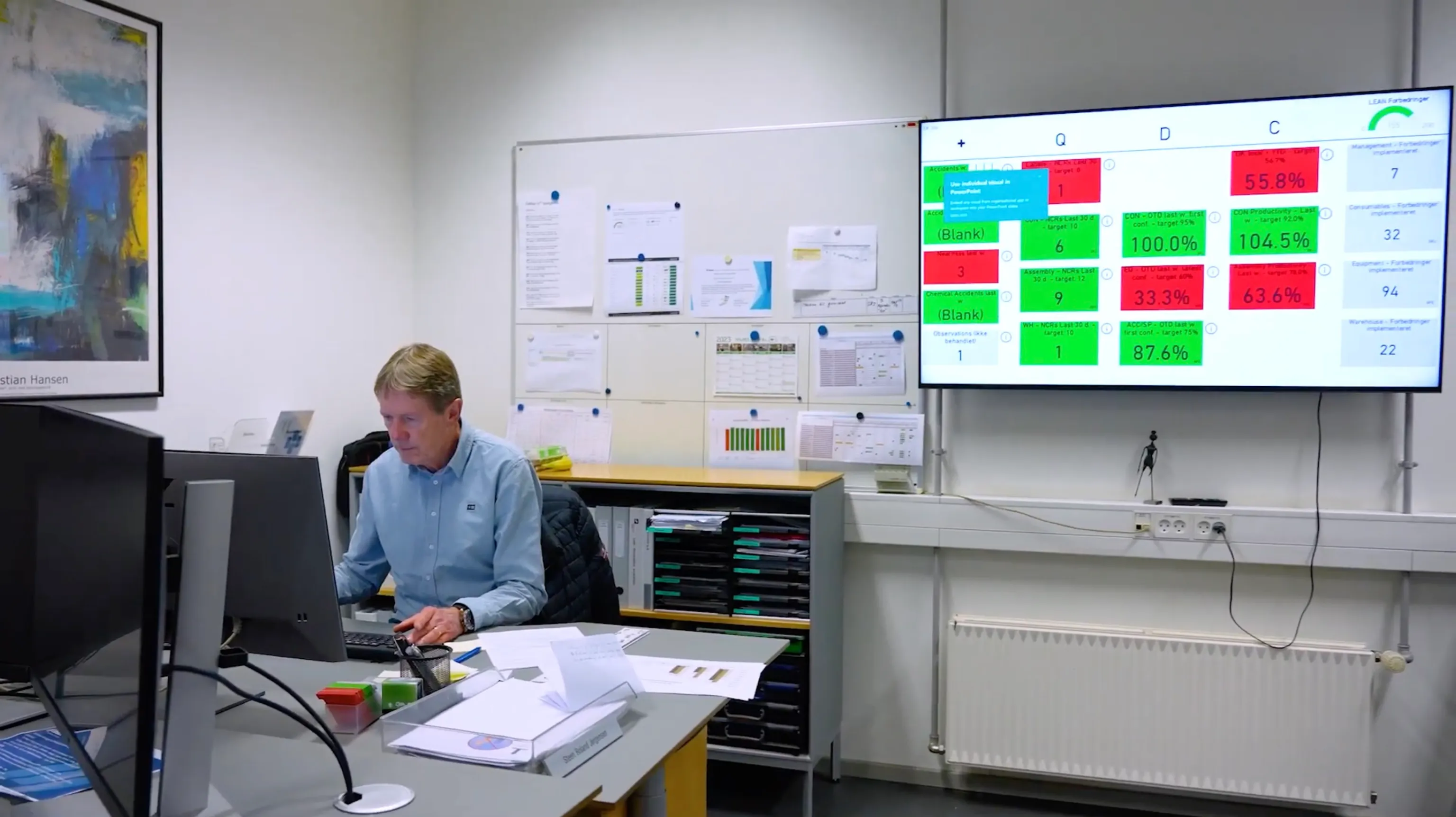 Production floor manager reviewing output, downtime, and yield metrics on a wall-mounted TV in a manufacturing environment