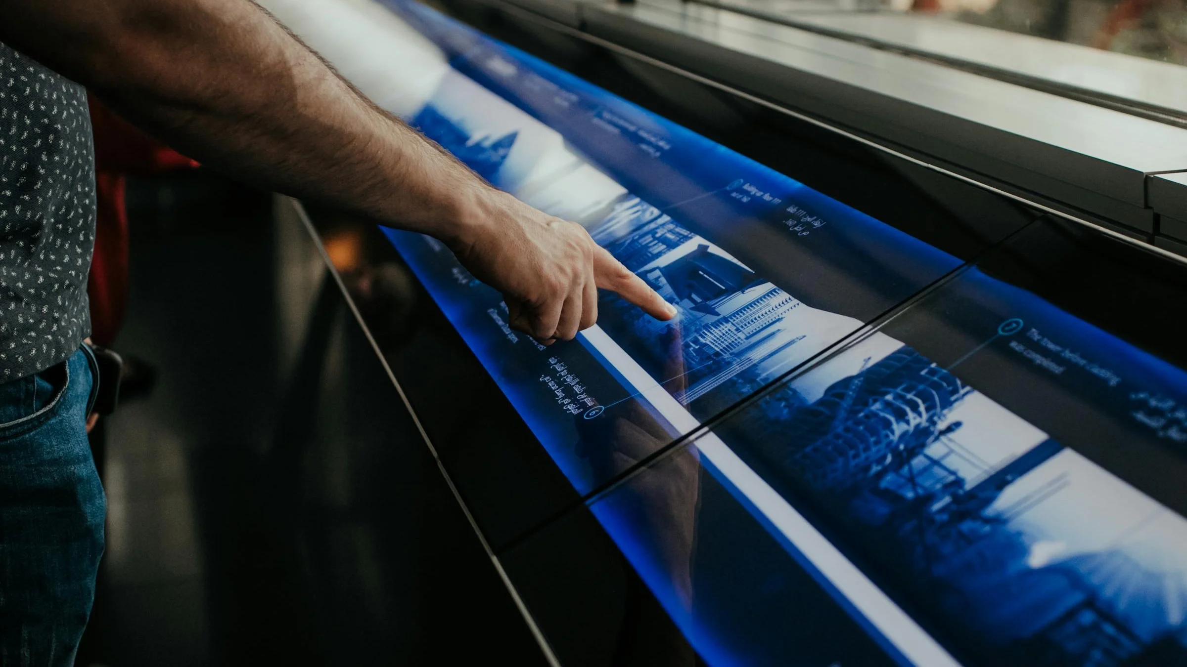 Close-up of a hand pressing an angled interactive touch table in an exhibition space, the blue-lit screen showing an illustrated city timeline
