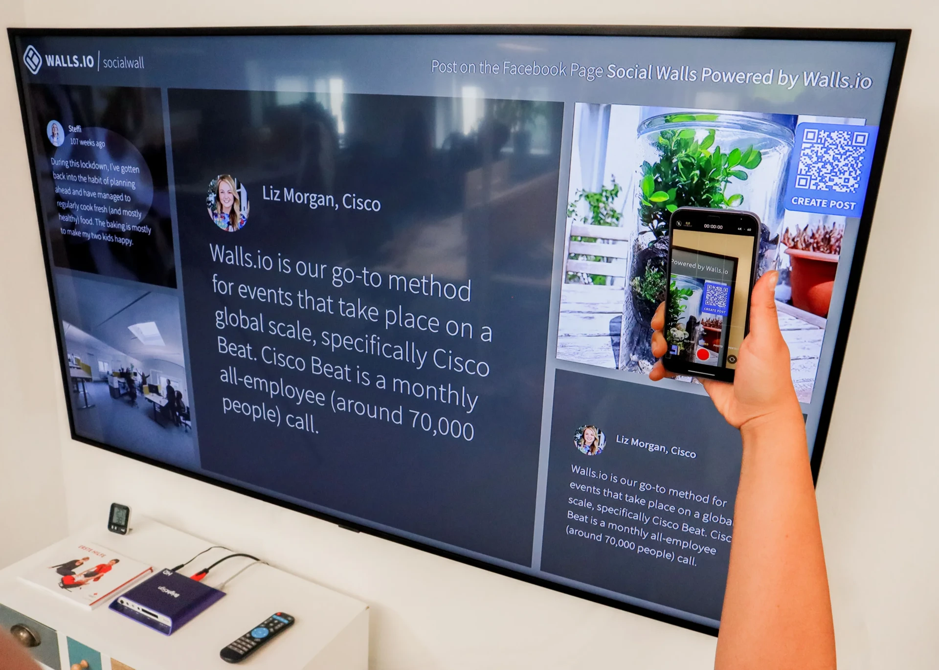 Wall-mounted TV showing a Walls.io social wall in a showcase layout featuring a Cisco testimonial alongside a QR code prompting viewers to post