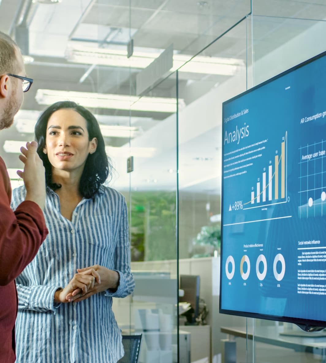 A woman and man are standing in front of an office digital signage screen that's displaying a TV dashboard, discussing the data on screen. UI elements of Fugo's digital signage solution are overlain on the photo.