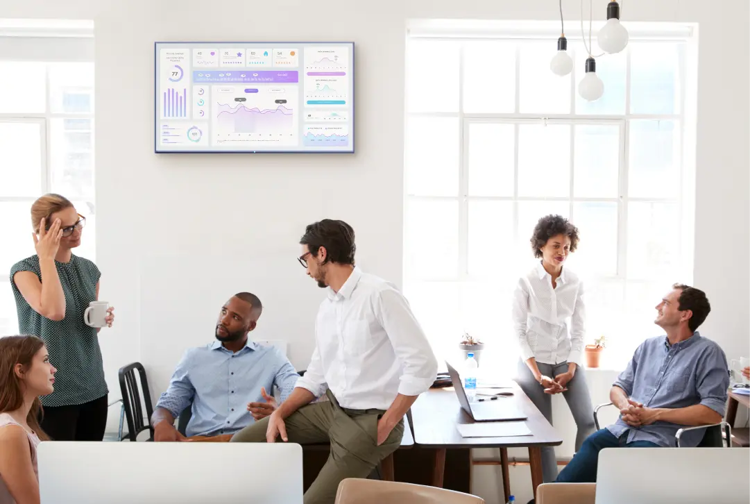 A TV dashboard is displayed on a screen on a white wall in an office. Employees are talking with each other.